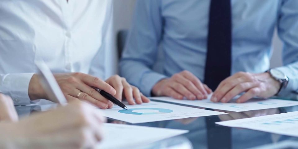 Three people in business attire sit at a glass table, reviewing documents with blue charts and graphs. One person points at a chart with a pen, indicating discussion or analysis.