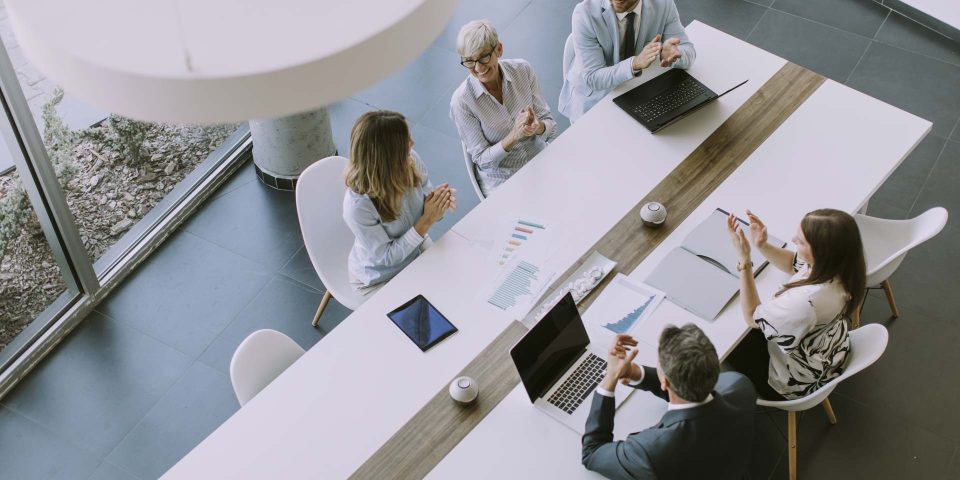 Five professionals sit around a white table in a modern office, discussing documents and charts. Laptops, tablets, and papers are on the table, and large windows provide natural light.